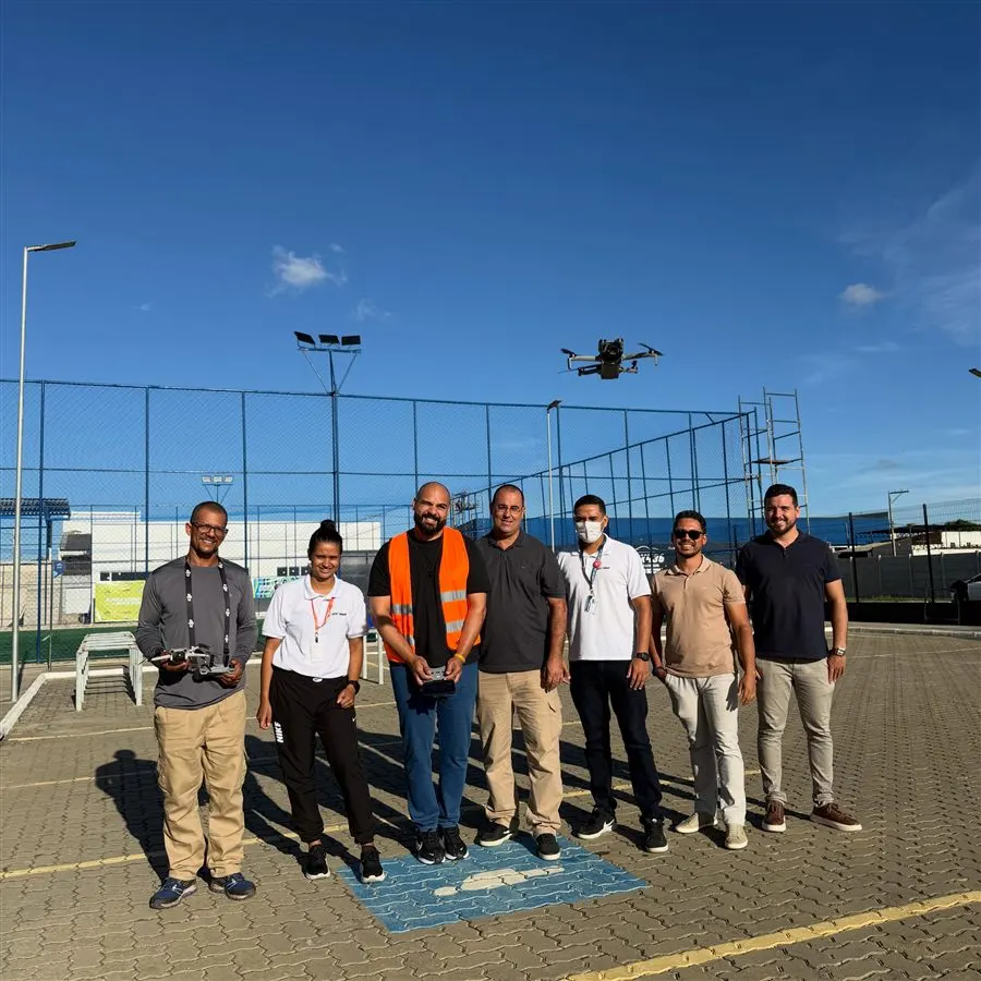 Grupo de alunos posando com os controles enquanto um drone sobrevoa a quadra